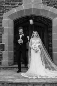 A bride in a wedding dress holds a bouquet before arched doors. Captured by top Wedding Photographers Toronto, the groom is blurred as he quickly steps down the stone stairs.