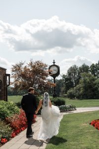 A bride and groom walk hand in hand on a path with flowers under a bright sky, passing a large outdoor clock and lush greenery, captured perfectly by Wedding Photographers Toronto.