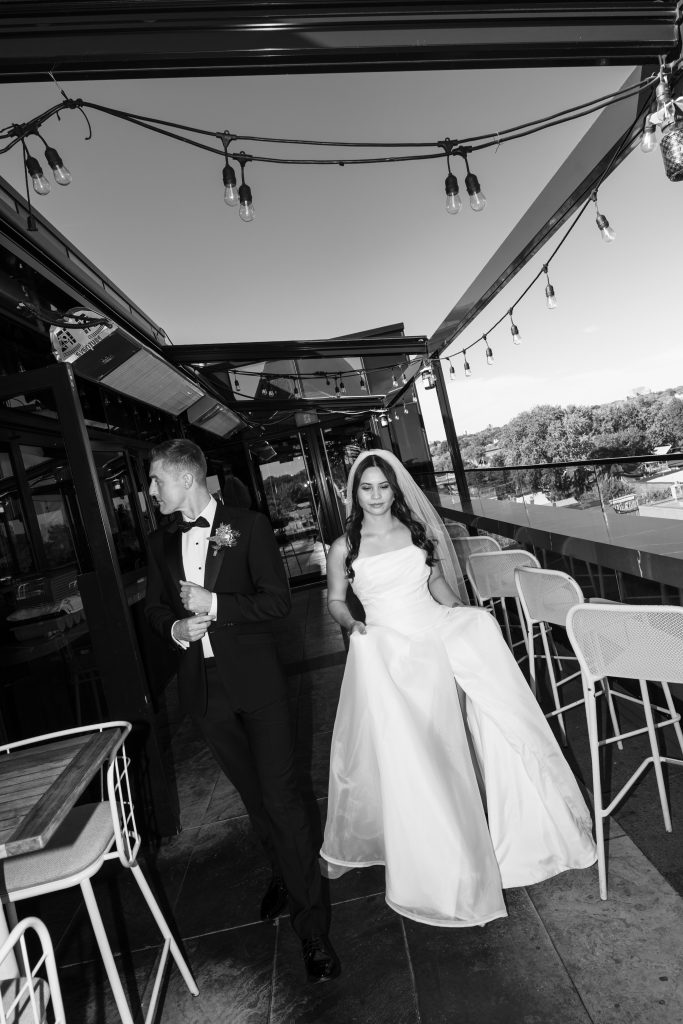 A bride in a white dress walks beside a groom in a tuxedo on an outdoor patio, both under string lights, with trees and sky in the background.
