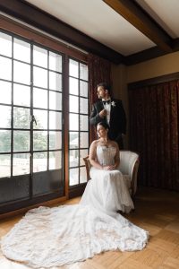 A bride in a white wedding dress sits on a chair while the groom, in a black tuxedo, stands behind her as they gaze out large windows—an elegant moment captured by Wedding Photographers Toronto.