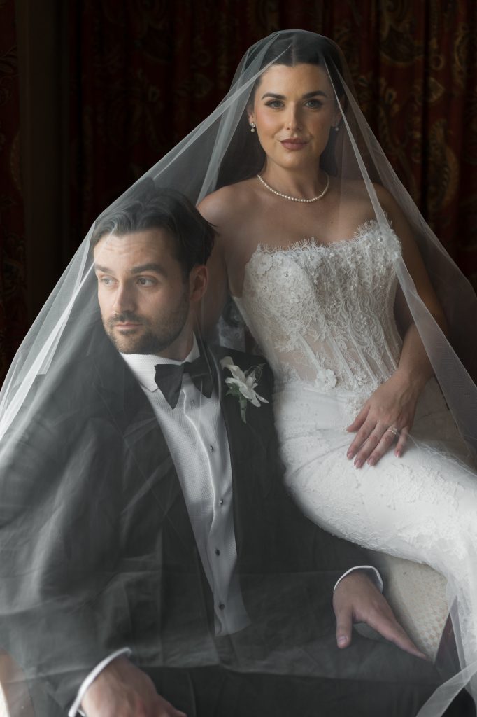 A bride in a lace wedding dress and groom in a tuxedo sit together under a sheer veil, looking in different directions, with elegant, soft lighting.
