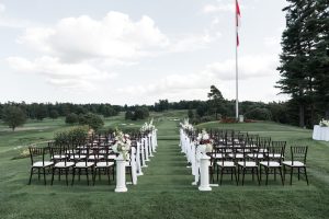 Rows of black chairs with white cushions are set up outdoors on grass for a wedding ceremony, perfect for Wedding Photographers Toronto to capture stunning moments, with flower arrangements lining the aisle and a large flagpole in the background.