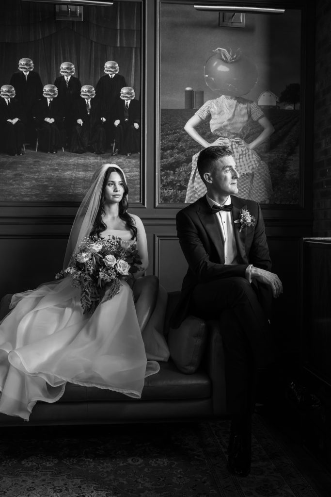 A bride and groom sit on a couch in formal attire, with the bride holding a bouquet. Behind them are surreal, artistic portraits on the wall. The image is in black and white.