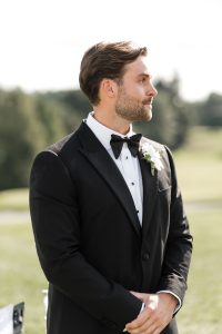 A man in a black tuxedo with a white shirt and black bow tie stands outdoors, looking to the side—an elegant moment captured by Wedding Photographers Toronto, with greenery and trees blurred in the background.