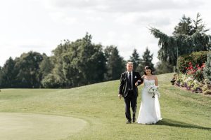 A bride in a white dress and an older man in a suit walk together on green grass, surrounded by trees and flowers, under a bright sky—captured beautifully by Wedding Photographers Toronto.