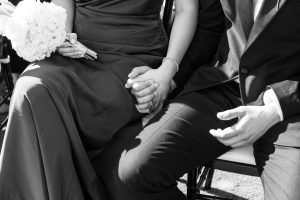 A couple dressed formally sits close together, holding hands. One holds a bouquet of flowers, while sunlight highlights their joined hands—perfectly captured by Wedding Photographers Toronto for a special occasion.