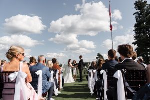 A bride and groom stand facing each other at an outdoor wedding ceremony, surrounded by guests under a partly cloudy sky. White ribbons decorate the chairs—a perfect scene for Wedding Photographers Toronto to capture.