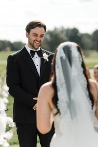A groom in a black tuxedo smiles at a bride in a white dress and veil during an outdoor wedding ceremony, beautifully captured by Wedding Photographers Toronto, with greenery visible in the background.