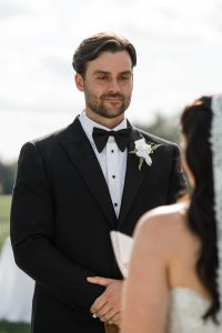 A man in a black tuxedo with a white boutonniere stands outdoors, facing a woman in a white dress and veil during what appears to be a wedding ceremony captured by Wedding Photographers Toronto.