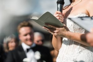 A bride in a white lace dress reads from a book and speaks into a microphone during an outdoor ceremony, beautifully captured by Wedding Photographers Toronto, with the groom and guests blurred in the background.