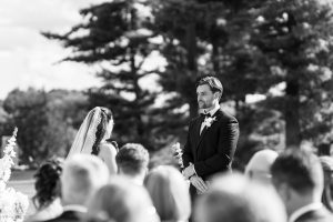 A bride and groom stand facing each other outdoors during their wedding ceremony, surrounded by guests. Tall trees fill the black and white scene—captured beautifully by Wedding Photographers Toronto.