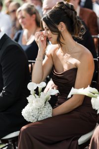 A woman in a brown dress sits holding a bouquet of white flowers, touching her face emotionally during an outdoor event captured by Wedding Photographers Toronto, with other people seated nearby.
