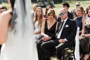 A seated man in a suit and sunglasses holds hands with a woman in a black dress as Wedding Photographers Toronto capture the couple watching the bride during an outdoor ceremony, with other guests observing in the background.