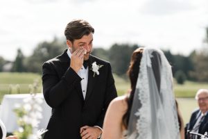 A groom wipes away a tear while facing his bride during an outdoor wedding ceremony, with greenery and guests visible in the background—captured perfectly by Wedding Photographers Toronto.