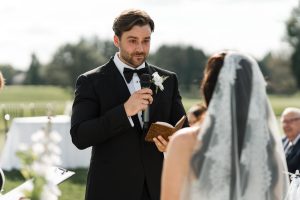 A groom in a black tuxedo holds a microphone and book, speaking to his bride in a white veil during an outdoor ceremony, perfectly captured by top Wedding Photographers Toronto.