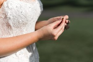 A bride in a white lace wedding dress holds a gold ring with both hands, standing outdoors on a sunny day—capturing moments like this is why Wedding Photographers Toronto are in such high demand.