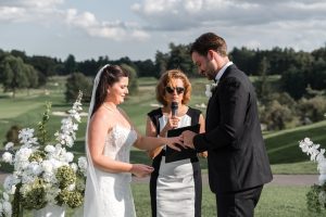 A bride and groom exchange rings outdoors during their wedding ceremony, with an officiant holding a microphone and white flowers in the background, beautifully captured by Wedding Photographers Toronto.