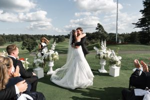 A bride and groom embrace and smile at an outdoor wedding ceremony, with Wedding Photographers Toronto capturing the moment amid applauding guests, white florals, a green golf course, and blue sky in the background.