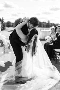 A groom dips and kisses his bride at an outdoor wedding ceremony, while guests nearby smile and applaud. Captured by Wedding Photographers Toronto, the black and white scene highlights her lace veil.