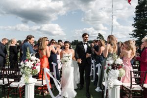 A bride and groom walk down the aisle outdoors, smiling and holding hands, as guests clap and celebrate. Wedding Photographers Toronto capture every joyful moment among beautiful flowers under a partly cloudy sky.