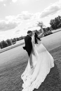 A bride and groom walk away on grass, the bride holding a bouquet up joyfully. Captured by Wedding Photographers Toronto, this black and white photo shows their wedding attire with trees and clouds in the background.