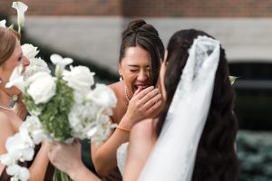 Three women in formal dresses, holding white flower bouquets. One woman laughs joyfully, covering her mouth, as another in a veil faces her—a candid moment captured by Wedding Photographers Toronto. The background is blurred.