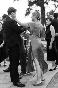 A woman in a dress smiles and touches a man’s face in an outdoor setting, surrounded by people in formal attire. Wedding Photographers Toronto capture this joyful, celebratory moment perfectly.