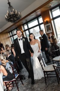 A bride in a white gown and a groom in a black tuxedo walk hand in hand, smiling, as Wedding Photographers Toronto capture their joyful moments at a reception with seated guests and large windows in the background.