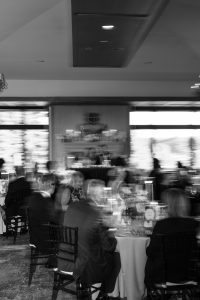 Black and white photo of people seated at round tables in a restaurant, with motion blur suggesting movement and conversation. Wedding Photographers Toronto capture the formal atmosphere enhanced by soft lighting and large windows.