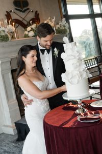 A bride and groom, dressed formally, smile as they cut a tall, white wedding cake with large floral decorations at an indoor reception—captured beautifully by Wedding Photographers Toronto.