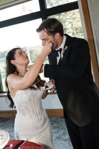 A bride in a white dress feeds cake to a groom in a black suit during their wedding reception, both smiling near a window with an outdoor view—captured beautifully by Wedding Photographers Toronto.