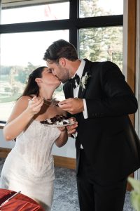 A bride and groom share a kiss as she holds a fork and a plate of cake, captured by Wedding Photographers Toronto. They stand in wedding attire near large windows with trees visible outside.