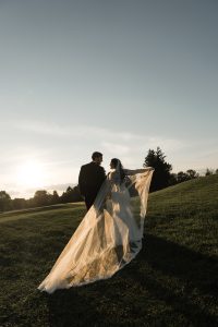 A bride and groom walk hand in hand on a grassy hill at sunset, her long veil trailing behind as sunlight casts a warm glow—captured beautifully by Wedding Photographers Toronto.