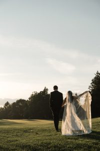 A bride and groom walk on a grassy field at sunset. The bride holds up her veil as Wedding Photographers Toronto capture the moment, with trees in the background under a clear sky.