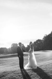 A bride and groom stand on a grassy field at sunset, holding hands and facing each other, as Wedding Photographers Toronto capture the moment with trees and a bright sky in the background.