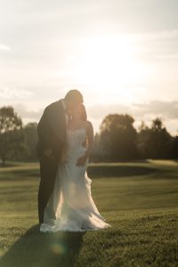A bride and groom stand closely together, sharing a kiss outdoors at sunset on a grassy field, with trees and soft sunlight in the background—beautifully captured by Wedding Photographers Toronto.