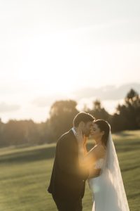 A bride and groom embrace outdoors at sunset, with soft sunlight creating a warm, romantic glow on the green lawn and trees—a perfect moment captured by Wedding Photographers Toronto.
