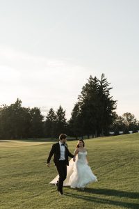 A bride and groom hold hands and walk across a grassy field at sunset, both smiling. Captured by Wedding Photographers Toronto, tall trees line the background under a clear sky.