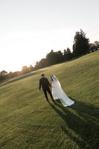 A bride and groom walk hand-in-hand across a sunlit grassy field at sunset, casting long shadows behind them—captured beautifully by Wedding Photographers Toronto.
