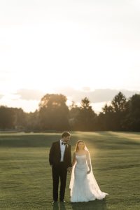A bride and groom stand hand-in-hand on a grassy field at sunset, with trees in the background and sunlight softly illuminating the scene—captured perfectly by Wedding Photographers Toronto.
