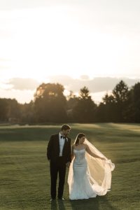A bride and groom walk hand in hand on a sunlit lawn, her veil flowing behind her and trees silhouetted in the background—captured beautifully by Wedding Photographers Toronto.