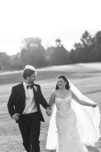 A bride and groom, smiling and holding hands, walk across a grassy field on their wedding day. Captured in black and white by Wedding Photographers Toronto, the moment is timeless and elegant.
