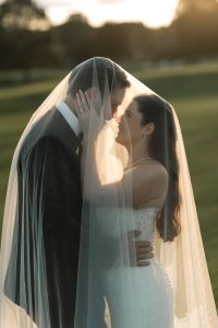 A bride and groom stand close together outdoors, smiling under a sheer wedding veil, bathed in warm sunlight—captured beautifully by Wedding Photographers Toronto.