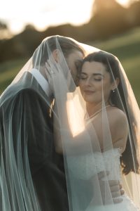 A bride and groom embrace tenderly under a sheer veil outdoors, bathed in soft, golden sunlight. Captured by top Wedding Photographers Toronto, the bride smiles as the groom gently kisses her temple.