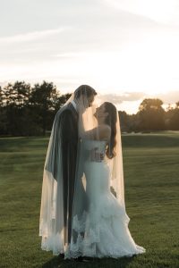 A bride and groom stand close together outdoors at sunset, wrapped in a wedding veil, sharing an intimate moment on a grassy field—captured beautifully by Wedding Photographers Toronto.