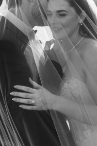 A bride and groom stand close together, partially covered by a flowing veil. Captured by Wedding Photographers Toronto, the bride smiles, showing her engagement ring, while the groom wears a suit in black and white.