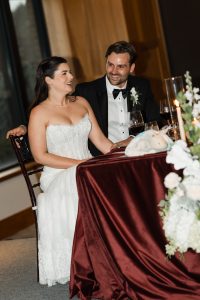 A bride and groom sit smiling together at a table with a burgundy tablecloth, floral arrangements, candles, and wine glasses—captured beautifully by Wedding Photographers Toronto during their wedding reception.
