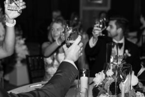 Black and white photo of people dressed formally, sitting at a table with flowers and candles, raising glasses in a toast—a joyful moment perfectly captured by top Wedding Photographers Toronto.