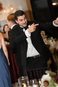 A man in a black tuxedo is smiling and pointing to the side at a formal event, with blurred guests and elegant decor—perfect inspiration for Wedding Photographers Toronto.