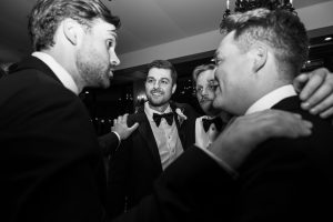 Four men in tuxedos stand close together, smiling and talking at what appears to be a formal event or wedding—captured beautifully by Wedding Photographers Toronto in classic black and white, showcasing camaraderie and celebration.